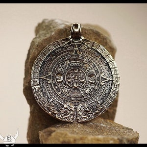 May include: A bronze pendant featuring a detailed replica of the Aztec Calendar Stone. The circular pendant depicts the stone's intricate carvings, including the central deity and surrounding glyphs.