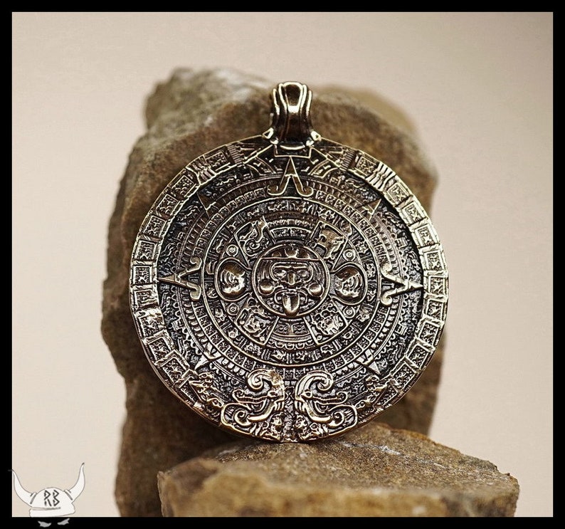 May include: A bronze pendant featuring a detailed replica of the Aztec Calendar Stone. The circular pendant depicts the stone's intricate carvings, including the central deity and surrounding glyphs.