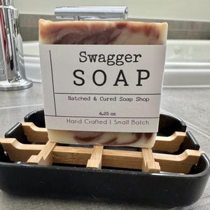 May include: A bar of brown and white swirl soap with a white label that reads "Swagger SOAP", "Batched & Cured Soap Shop", "4.25 oz", and "Hand Crafted | Small Batch". The soap is sitting on a wooden soap dish.