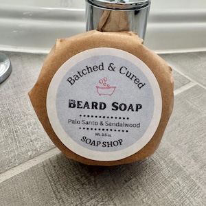 May include: A round beard soap wrapped in brown paper, with a label that reads "Batched & Cured BEARD SOAP Palo Santo & Sandalwood Wt. 3.5 oz SOAP SHOP". The soap is sitting on a tiled surface.