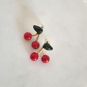 Può includere: Un paio di orecchini a lobo a forma di ciliegia con due sfere rosse lucide attaccate a un gambo dorato con una foglia verde scuro. Gli orecchini sono dotati di perno e chiusure in plastica trasparente per un fissaggio sicuro.