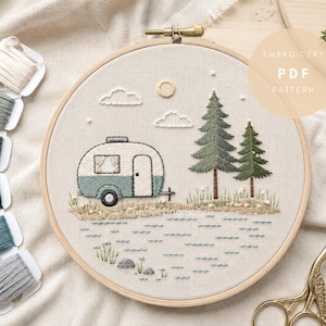 May include: An embroidery hoop containing a fabric panel with a stitched scene of a vintage camper, two pine trees, clouds, and a sun. The text EMBROIDERY PDF PATTERN is visible. Six spools of embroidery floss and gold scissors are included.