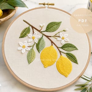 May include: A circular wooden embroidery hoop containing a fabric panel with a hand-stitched design of a lemon branch. The embroidery features two bright yellow lemons, green leaves, and white blossoms with yellow centers. Text reads EMBROIDERY PDF PATTERN.
