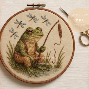 May include: An embroidered frog in overalls sits by the water, holding a fishing rod. The frog is green and brown, with detailed stitching. Dragonflies are embroidered above the frog. The embroidery is in a wooden hoop.