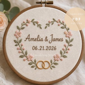 Könnte beinhalten: Ein Stickrahmen mit cremefarbenem Stoff und einem herzförmigen Blumenkranz in Rosa und Grün. In der Mitte befinden sich der Text Amelia & James und das Datum 21.06.2026 in braunem Garn, sowie zwei goldene Ringe am unteren Rand.