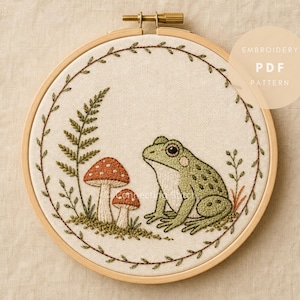Puede incluir: Un bastidor de bordado con una pieza de tela que presenta un diseño bordado de una rana verde, dos setas de sombrero rojo y una hoja de helecho, enmarcado por un borde circular de enredadera. El texto EMBROIDERY PDF PATTERN es visible.