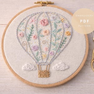 Puede incluir: Un bastidor de bordado redondo de madera que contiene un panel de tela con un diseño de globo aerostático cosido a mano. El globo presenta bordados florales en tonos pastel, enredaderas verdes y una cesta tejida. Dos nubes blancas bordadas. Tijeras de cigüeña doradas.