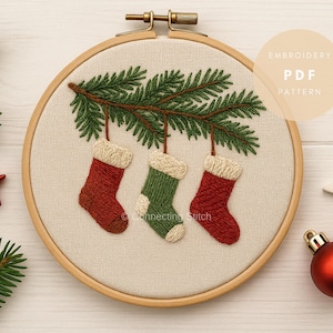May include: Embroidered Christmas stocking design in a wooden hoop. The design features three stockings in red and green, hanging from a green pine branch. The text "Embroidery PDF Pattern" is visible. Additional Christmas decorations are in the background.