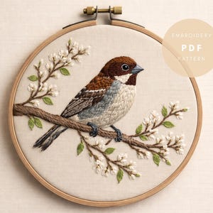 Può includere: Uccello ricamato su un ramo con fiori bianchi, in un telaio da ricamo in legno. L'uccello ha piume marroni, bianche e grigie. Il testo "EMBROIDERY PDF PATTERN" è in un cerchio a destra.
