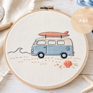 May include: An embroidery hoop containing a fabric panel with a stitched design of a light blue and cream-colored van with a coral surfboard on its roof. The scene includes a small stitched wave and a coral seashell on a sandy beach background.