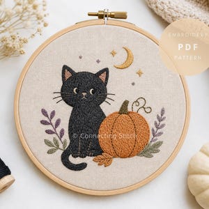 Peut inclure: Un tambour à broder contenant un tissu beige avec un motif brodé d'un chat noir assis à côté d'une citrouille orange. La scène comprend un croissant de lune jaune, de petites étoiles et un feuillage violet et vert. Texte EMBROIDERY PDF PATTERN.