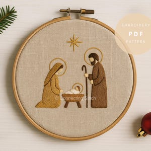 Könnte beinhalten: Gestickte Krippenszene in einem Holzreifen. Das Design zeigt Maria, Josef und das Jesuskind in einer Krippe, mit einem Stern darüber. Der Text "Embroidery PDF Pattern" ist sichtbar. Ein Tannenzapfen und eine rote Christbaumkugel sind ebenfalls vorhanden.