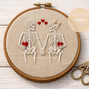 May include: Embroidered hoop art featuring two skeleton figures holding hands, each holding a red heart. The skeletons are white against a tan fabric background, framed by a wooden hoop. A small bow adorns one skeleton. Gold scissors are in the corner.