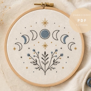 May include: An embroidery hoop containing a white fabric panel with a stitched design of moon phases in blue and black thread, accented by gold stars and a central floral motif. The text EMBROIDERY PDF PATTERN is visible in the upper right corner.