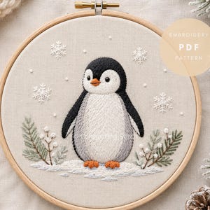 Op de afbeelding: Een ronde houten borduurring met een stoffen paneel met een handgeborduurd pinguïnontwerp. De pinguïn is zwart, wit en grijs met oranje voeten, staat op sneeuw en is omringd door witte sneeuwvlokken en groene dennentakken. Tekst: EMBROIDERY PDF PATTERN.