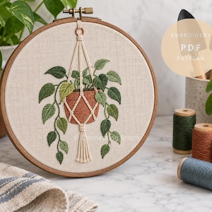 Può includere: Un telaio da ricamo rotondo in legno contenente un pannello in tessuto con il disegno ricamato di una pianta sospesa in un vaso di terracotta tenuto da un cordino in macramè. Il testo EMBROIDERY PDF PATTERN è visibile in alto a destra.