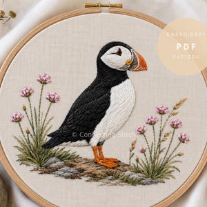 May include: An embroidery hoop containing a fabric panel with a stitched puffin bird in black, white, and orange, surrounded by pink flowers and green grass. A circular overlay in the top right corner displays the text EMBROIDERY PDF PATTERN.