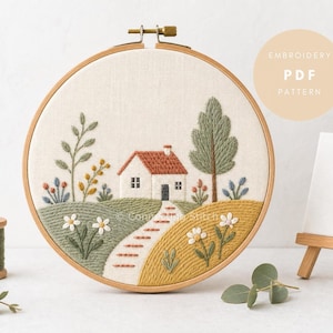 Pode incluir: Um bastidor de bordado contendo um painel de tecido com uma paisagem bordada apresentando uma casa branca com telhado vermelho, uma árvore verde alta, colinas em tons de verde e amarelo, um caminho e flores. O texto diz EMBROIDERY PDF PATTERN.