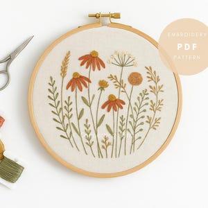Può includere: Un telaio da ricamo con un motivo floreale con fiori arancioni e gialli e foglie verdi. Il testo "EMBROIDERY PDF PATTERN" è in alto a destra. Forbici e rocchetti di filo arancione, giallo e verde a sinistra.