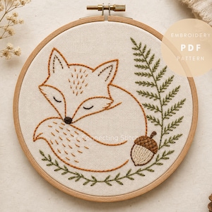 May include: A circular wooden embroidery hoop containing a cream-colored fabric with a hand-stitched design of a sleeping fox, a green fern frond, and a brown acorn. The text EMBROIDERY PDF PATTERN is visible in the upper right corner of the image.