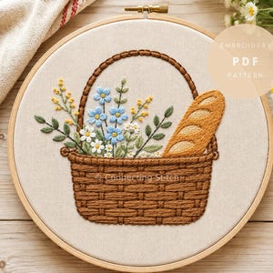 May include: An embroidery hoop containing a fabric panel with a stitched design of a brown woven basket filled with blue and white flowers, green leaves, and a golden-brown baguette. The text EMBROIDERY PDF PATTERN is visible in the upper right.