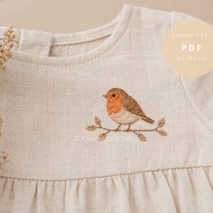 May include: A beige linen dress with an embroidered robin perched on a branch. The bird has a red breast and brown wings. The dress has a round neckline and short sleeves. Text on the image reads "Embroidery PDF Pattern".