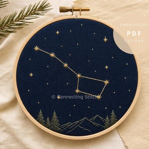 May include: A circular wooden embroidery hoop containing dark blue fabric with gold thread stitching depicting the Big Dipper constellation, scattered stars, and a mountain range with pine trees at the bottom. Text reads EMBROIDERY PDF PATTERN.