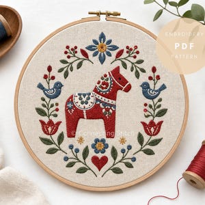 May include: A circular wooden embroidery hoop containing a beige fabric with a hand-stitched design of a red Dala horse centered within a floral wreath. The wreath features blue birds, red flowers, and green leaves. Text reads EMBROIDERY PDF PATTERN.