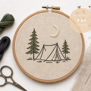 May include: A circular wooden embroidery hoop containing a beige fabric panel with a stitched design of a tent, two pine trees, and a crescent moon. The text EMBROIDERY PDF PATTERN is visible in a circular overlay. Includes olive and dark gray thread.