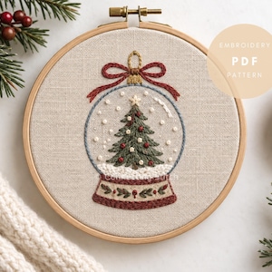 May include: An embroidery hoop containing a beige fabric panel with a hand-stitched snow globe design. The globe features a green Christmas tree with red and white ornaments, a red bow at the top, and a base with green leaves and red berries.