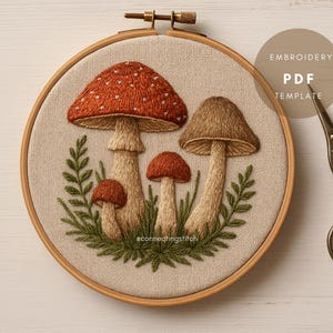 Könnte beinhalten: Gesticktes Pilzdesign in einem Holzreifen. Die Pilze haben braune und rote Kappen mit weißen Punkten, umgeben von grünem Laub. Der Text "EMBROIDERY PDF TEMPLATE" ist sichtbar.
