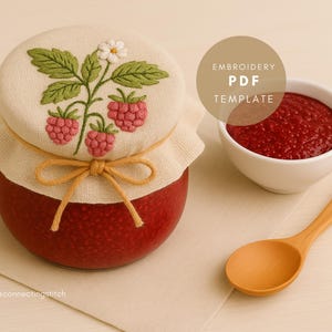 Puede incluir: Un tarro rojo con una tapa crema bordada con un diseño de frambuesas. Un pequeño cuenco blanco con mermelada roja y una cuchara de madera están junto al tarro. La tapa está atada con una cuerda beige.