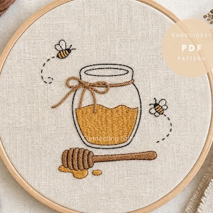 Puede incluir: Un bastidor de bordado circular de madera que contiene una tela de lino con un diseño bordado a mano de un tarro de cristal lleno de miel dorada, atado con un lazo de cuerda marrón, rodeado por dos pequeñas abejas y un cazo de madera.