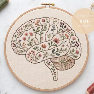 以下が含まれることがあります： クリーム色の布が入った刺繍枠で、人間の脳の手刺繍デザインが施されています。脳は暗い色の糸で輪郭が描かれ、様々な小さくカラフルな花や植物の模様で満たされています。テキストには「EMBROIDERY PDF PATTERN」と書かれています。