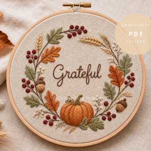 May include: A circular wooden embroidery hoop containing a linen fabric piece with a hand-stitched autumn wreath design. The embroidery features a central orange pumpkin, brown acorns, red berries, wheat stalks, and green and orange oak leaves with the word Grateful stitched in brown thread.