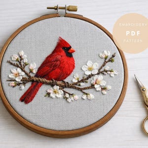 May include: Embroidered cardinal bird on a branch with white flowers, set in a wooden hoop. The bird is red with a black mask. The embroidery is on a gray fabric. The text "Embroidery PDF Pattern" is in a circle.