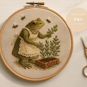 May include: An embroidery hoop with a detailed frog wearing an apron, tending to a small garden. The frog is green, and the apron is beige with a lace trim. Bees are embroidered above the frog. The text "Embroidery PDF Pattern" is in a circle.
