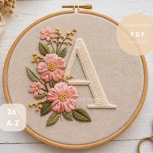 Puede incluir: Letra "A" bordada en un aro de madera, adornada con flores rosas y hojas verdes. El diseño incluye el texto "Embroidery PDF Pattern" y "26 A-Z". Un par de tijeras doradas descansa cerca.