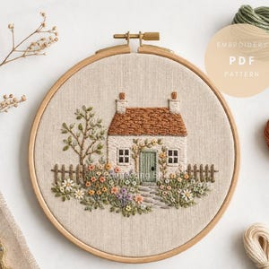 May include: A round wooden embroidery hoop containing a fabric panel with a stitched scene of a cottage with a brown roof, a green door, and a stone path surrounded by colorful flowers, a tree, and a fence, with the text EMBROIDERY PDF PATTERN.