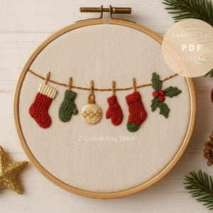 May include: Embroidered Christmas hoop art featuring miniature stockings, mittens, and ornaments hanging on a string. The design includes a holly sprig and is set against a cream-colored fabric within a wooden hoop. The text "Embroidery PDF Pattern" is visible.