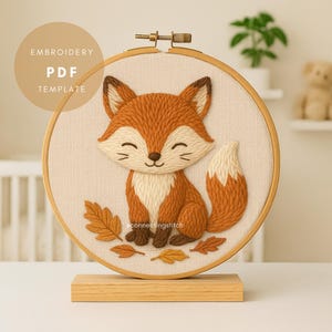 Può includere: Un telaio da ricamo con una volpe sorridente con pelliccia arancione e bianca, circondata da foglie autunnali. Il testo "EMBROIDERY PDF TEMPLATE" è in un cerchio sopra la volpe. Il ricamo è montato su un supporto di legno.