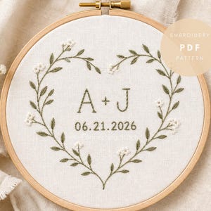 May include: A circular wooden embroidery hoop containing white fabric with a hand-stitched heart-shaped wreath of green leaves and small white flowers. The center features the embroidered letters A + J and the date 06.21.2026 in dark green thread.