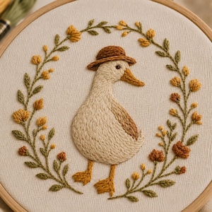 Peut inclure: Un tambour à broder circulaire contenant un panneau en tissu avec un motif brodé à la main représentant un canard crème portant un chapeau marron, centré dans une couronne de fleurs jaunes et orange avec des feuilles et tiges vertes.