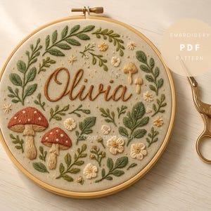 May include: Embroidered hoop art featuring the name "Olivia" in brown thread, surrounded by green leaves, mushrooms, and flowers. The design is set against a beige background within a wooden hoop. A pair of gold scissors rests nearby.