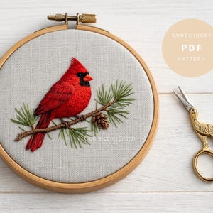 May include: An embroidery hoop featuring a red cardinal bird on a branch with pine needles and a pinecone. The embroidery is on light grey fabric. Gold scissors and a circular graphic with text are also in the image.