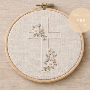 Pode incluir: Um bastidor de bordado com tecido bege com um desenho de cruz. A cruz é adornada com flores bordadas em branco, rosa e azul. O texto "EMBROIDERY PDF PATTERN" está em um círculo bege.