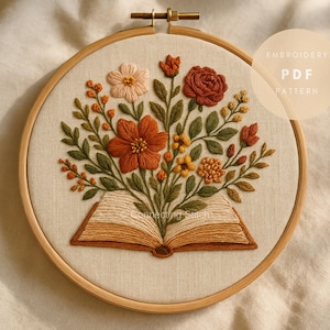 Può includere: Design floreale ricamato in un cerchio di legno. L'opera presenta un bouquet di fiori nei toni dell'arancione, pesca e giallo, che esce da un libro aperto. Il testo "EMBROIDERY PDF PATTERN" è visibile.