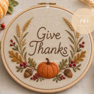 May include: A circular wooden embroidery hoop containing beige fabric with the stitched text Give Thanks. The design features an orange pumpkin, brown acorns, red berries, green leaves, and golden wheat stalks arranged in a decorative wreath pattern.
