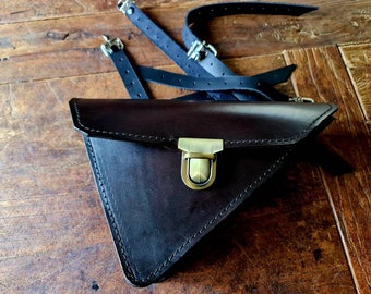 May include: A dark brown leather triangle-shaped bag with a brass clasp. The bag has a stitched edge and is lying on a wooden surface.