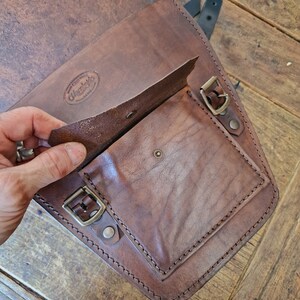 May include: A brown leather motorcycle saddlebag with a flap that opens to reveal a smaller pocket. The bag has a brass buckle and stitching around the edges. The bag has a leather label that says "Wayward" with a small logo.